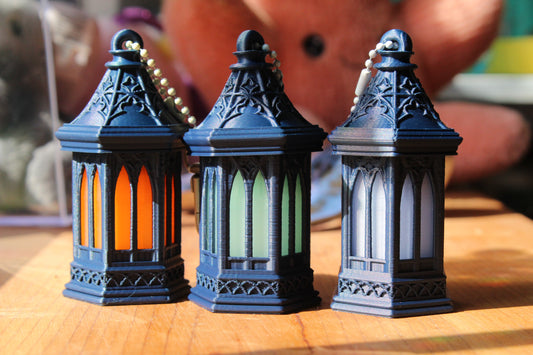 Three miniature gothic-style lanterns with dark blue-gray metal bodies and colorful illuminated glass panels, arranged side by side on a wooden surface.