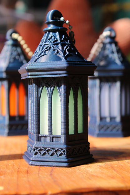 A close-up of a small decorative lantern with a dark blue-gray body and pale green glass panels, set on a wooden surface with other similar lanterns blurred in the background.