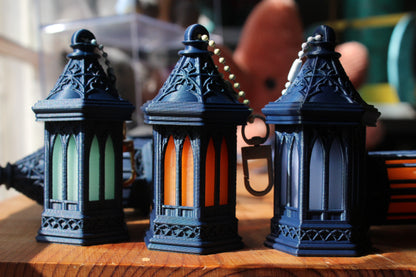Three miniature gothic-style lanterns with dark blue bodies and arched stained-glass windows in green, orange, and white, displayed on a wooden surface.