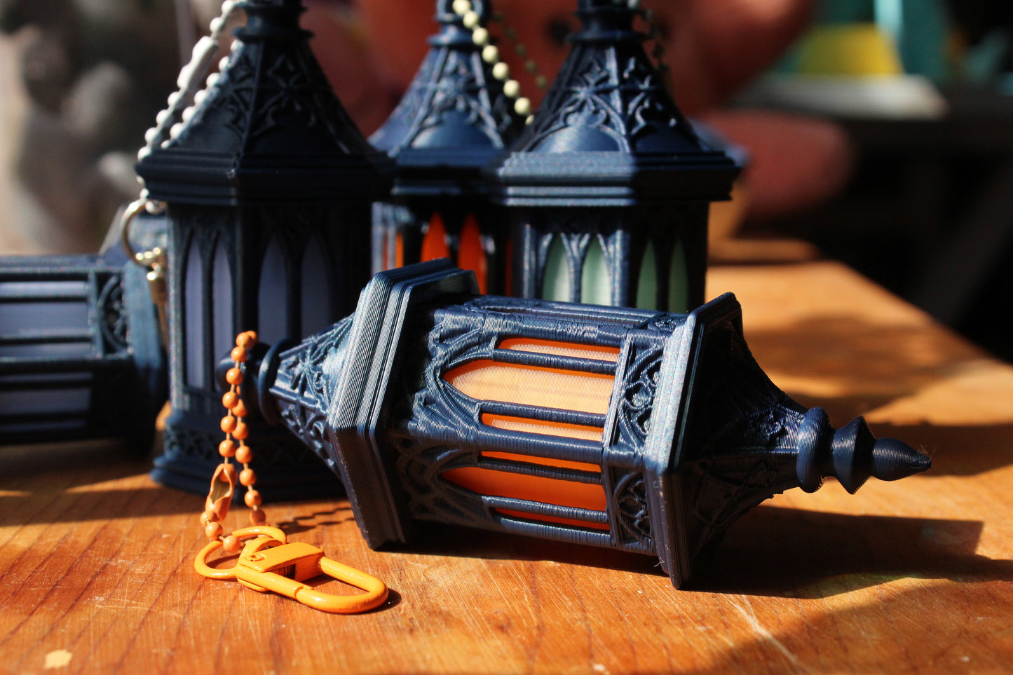 A close-up of a black ornate lantern with a gold-colored chain lying on a wooden surface, with other similar lanterns blurred in the background.