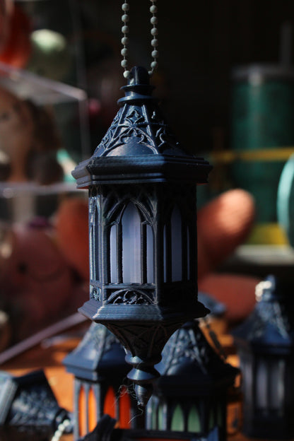 A close-up of a black decorative hanging lantern with ornate carved details and a ball chain pull, shown against a blurred background.
