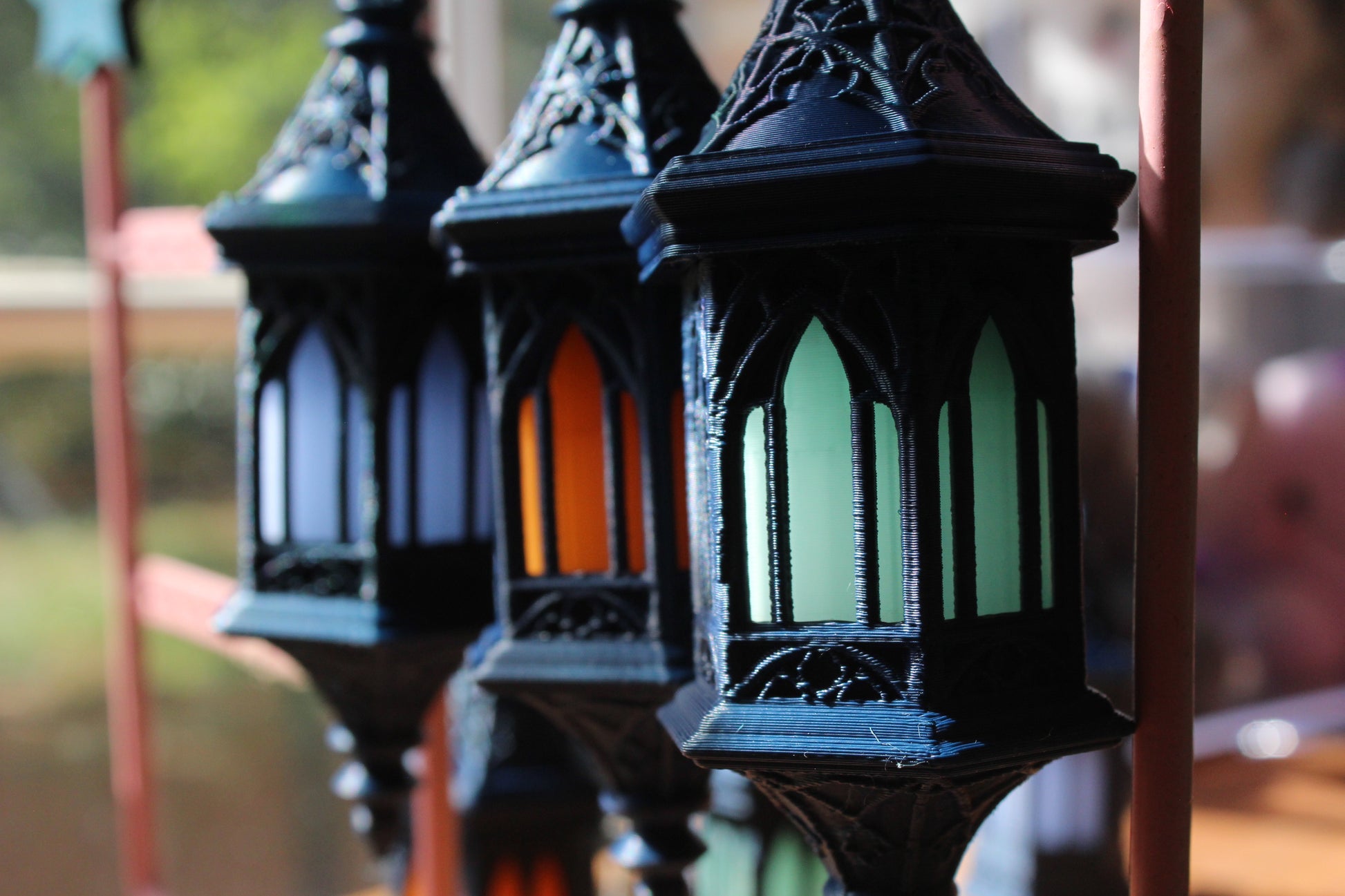 Three ornate black metal lanterns with arched stained-glass windows in blue, orange, and green, shown in a close-up outdoor shot with a blurred background.