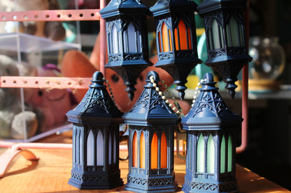 A set of decorative lanterns with dark blue-gray metal frames and colorful arched glass panels in orange, white, and green, displayed on a wooden surface.