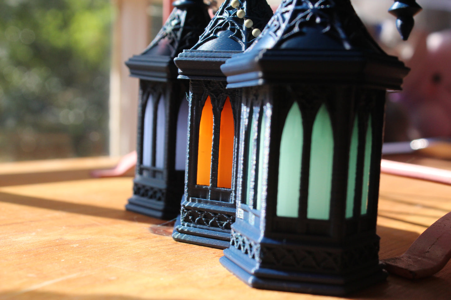 Three ornate black metal lanterns with arched glass panels, one glowing with orange light and two with pale green light, arranged on a wooden surface in warm natural light.